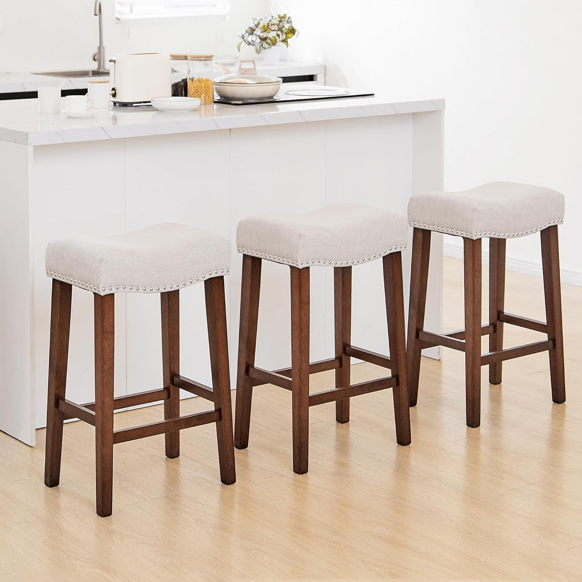 Three beige upholstered backless stools with wood legs lined in front of a white kitchen island.