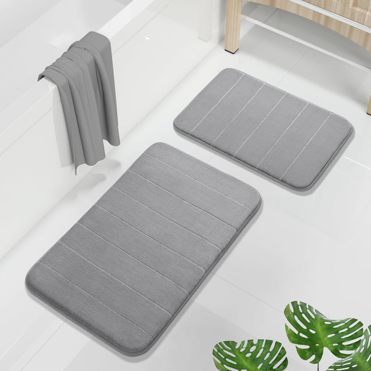 Two rectangular gray memory-foam bath mats on a glossy white tiled floor next to a tub and wooden stool.