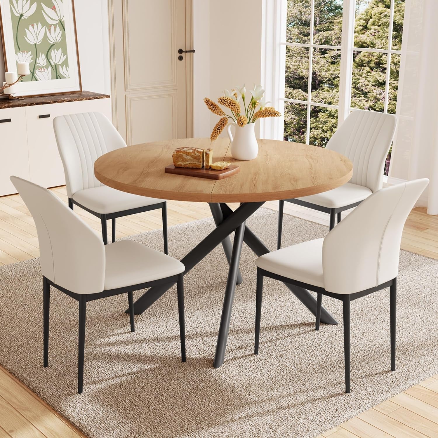 Round wood dining table with four cream upholstered U‑shaped chairs in a sunlit room