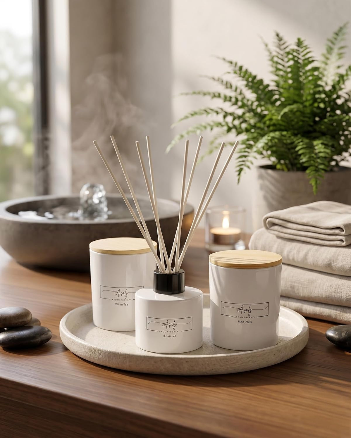 Styled lifestyle scene of two candles with wooden lids and reed diffuser arranged on a tray near towels and a plant.