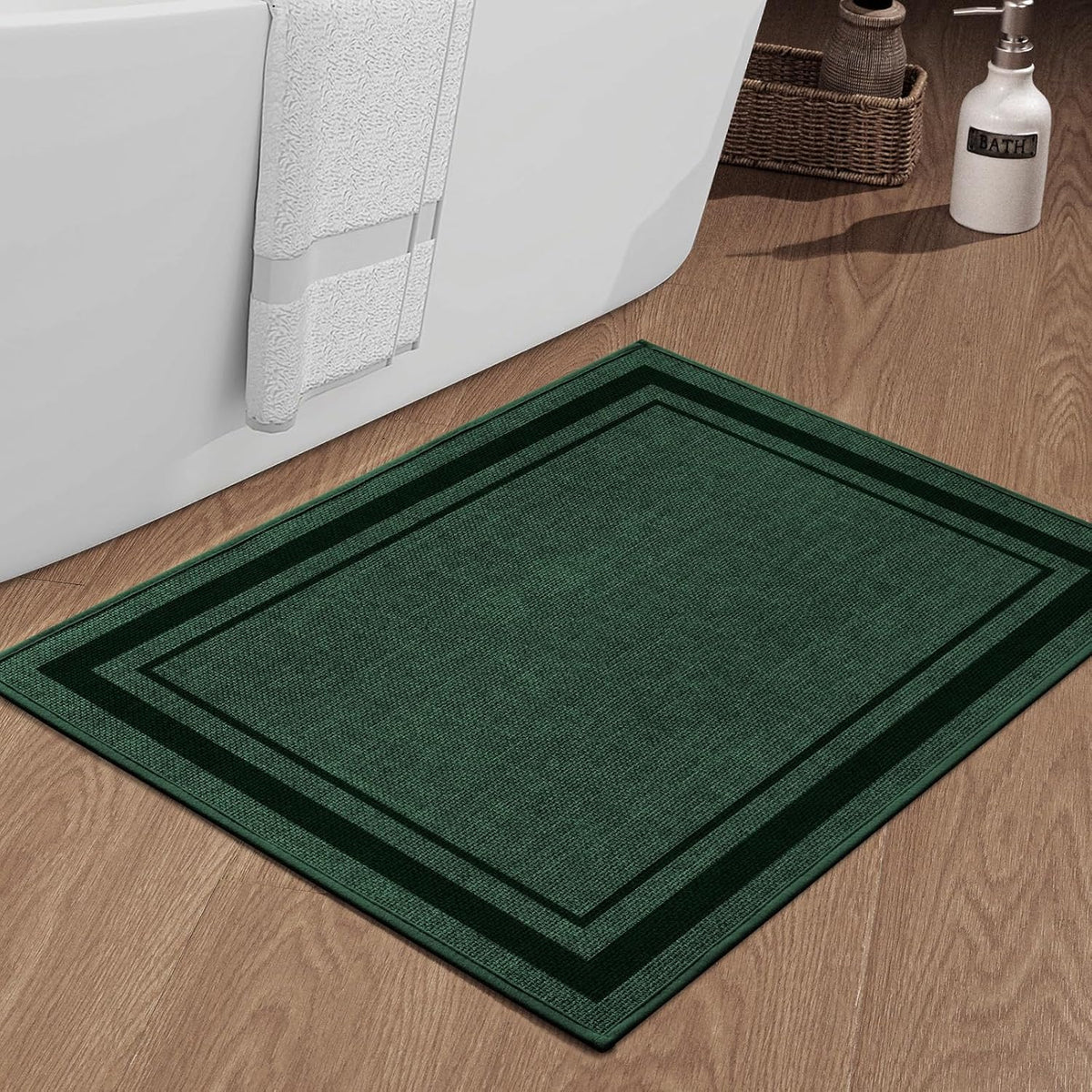 Rectangular dark green bath mat placed on wood floor beside a bathtub with a towel hanging on tub edge.