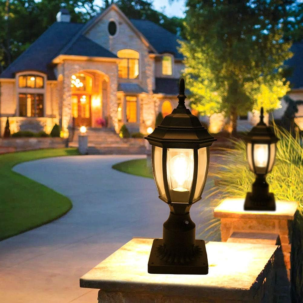 Evening driveway scene showing two lit post lanterns mounted on stone piers in front of a large house.
