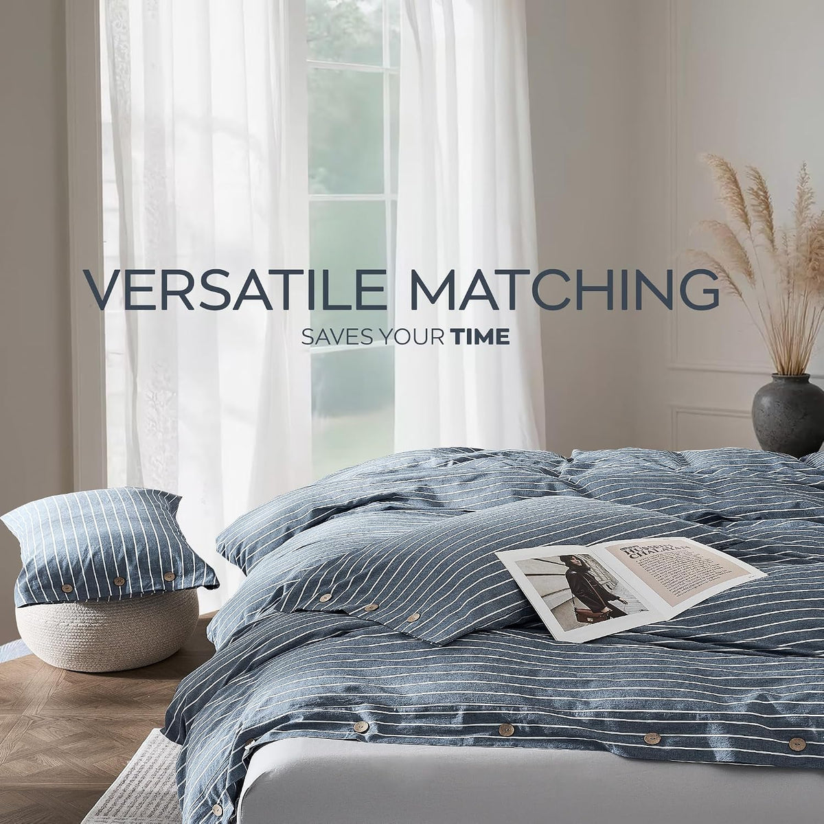 Blue striped duvet and pillows on a made bed in front of sheer curtains with a side vase on a table.