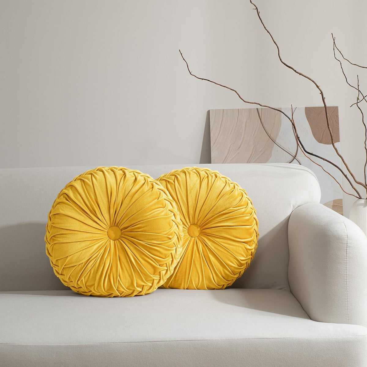 Two round ruched yellow pillows on a light-gray sofa with branches and abstract art behind.