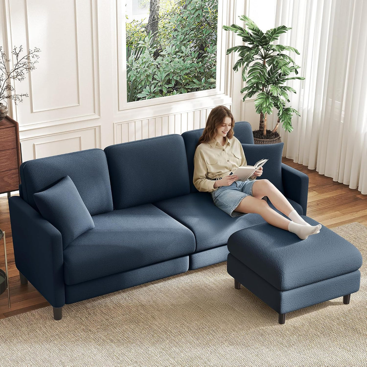 Navy blue loveseat with separate ottoman, woman seated and reading in a bright room.