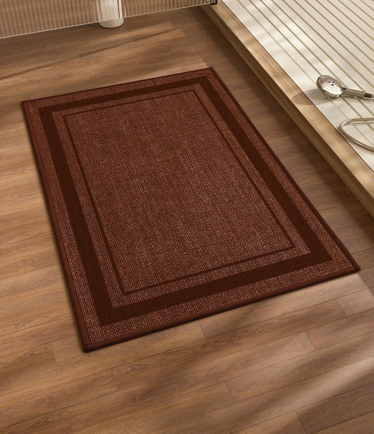 Rectangular brown bath mat with double border placed on wood floor beside a tub platform