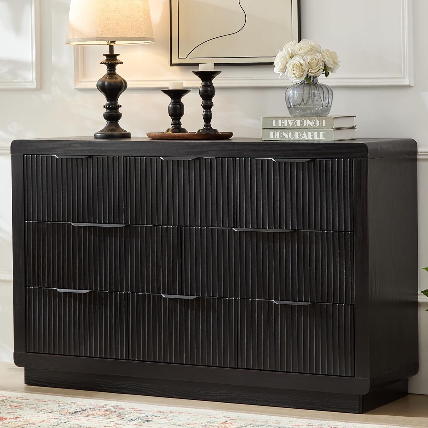 Wide black ribbed-front dresser with six drawer fronts, topped with a lamp, candles and a small stack of books.