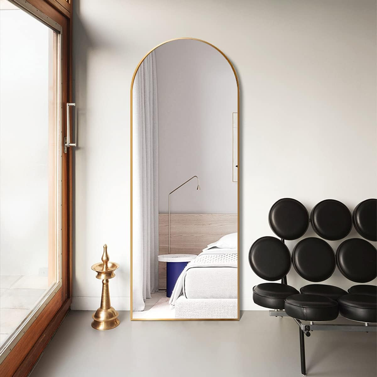 Tall arched gold mirror standing on floor beside a black circular-segment chair and a windowed doorway.