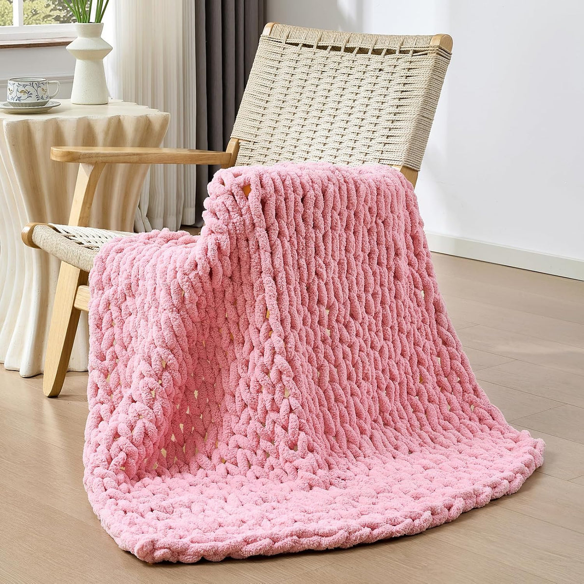 Soft pink chunky knit throw casually draped over a woven chair with a side table holding a teacup.