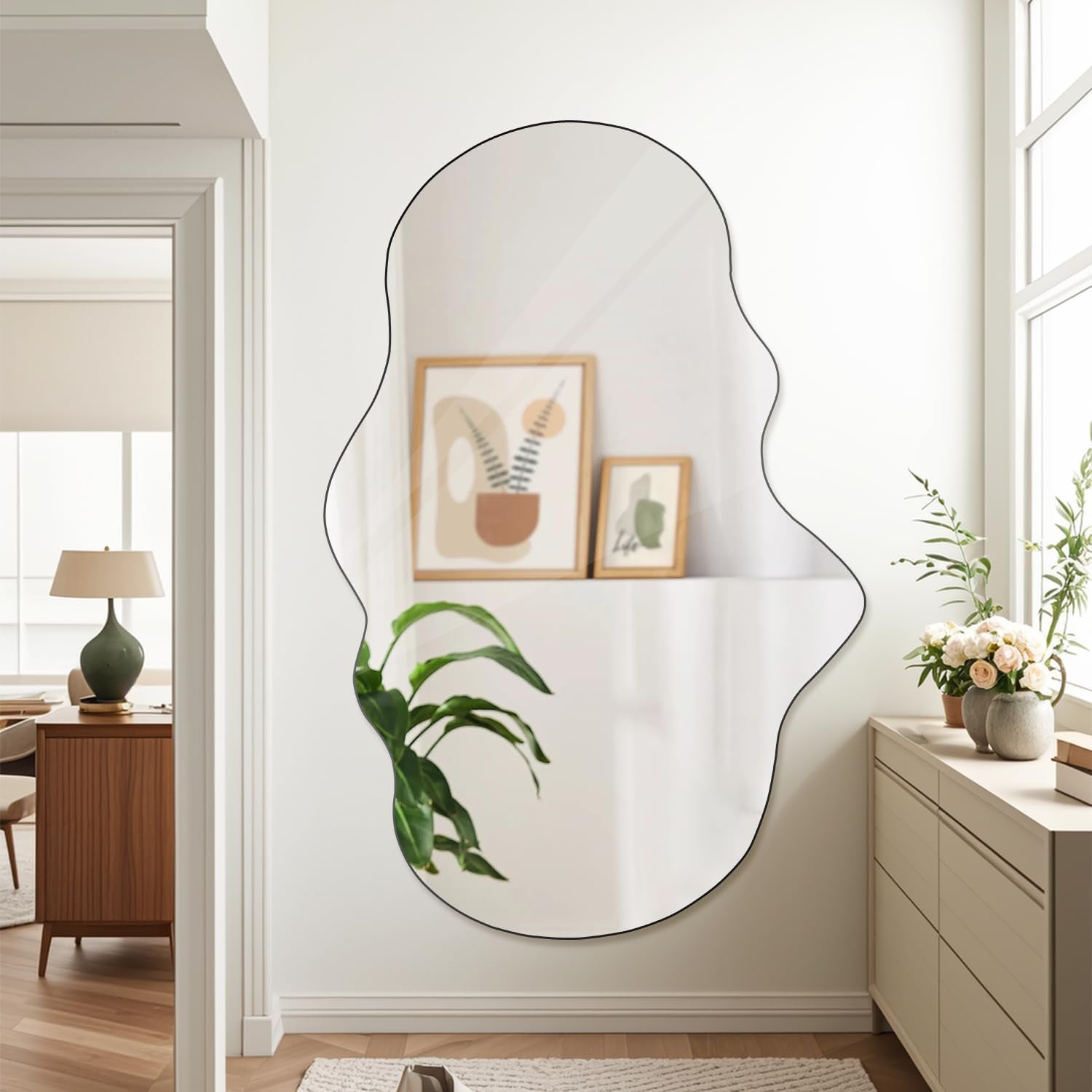 Large irregular frameless mirror mounted on a white wall reflecting framed artwork and a nearby houseplant in a hallway