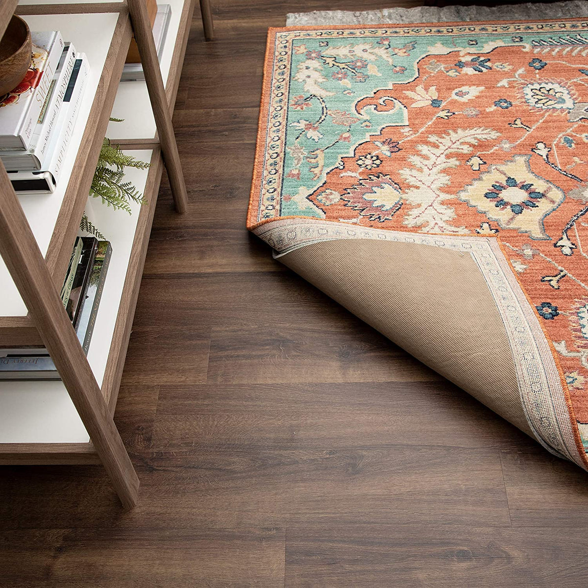 Side view of a rug corner lifted to show a smooth tan backing beneath the patterned rug on a wood floor.