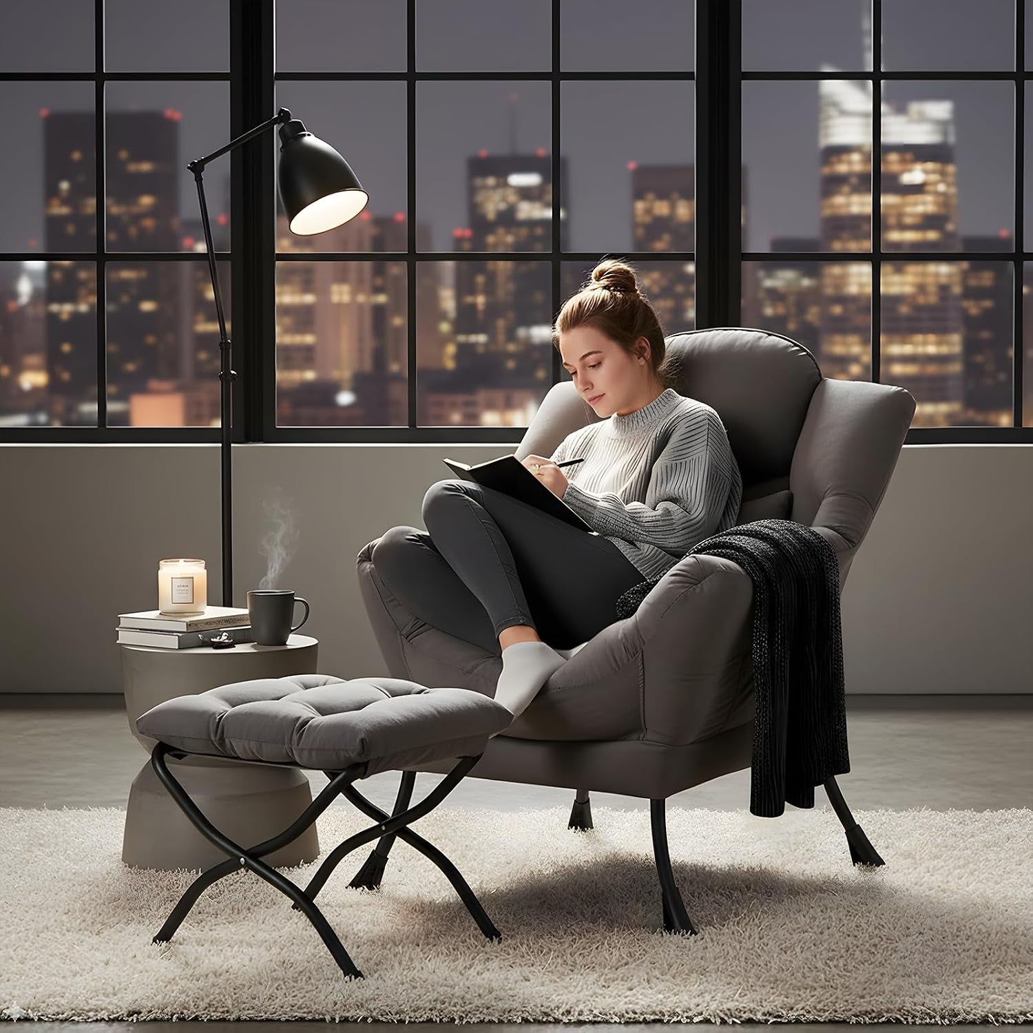 Person writing in a notebook while seated in the gray chair with ottoman in a city-view room at night