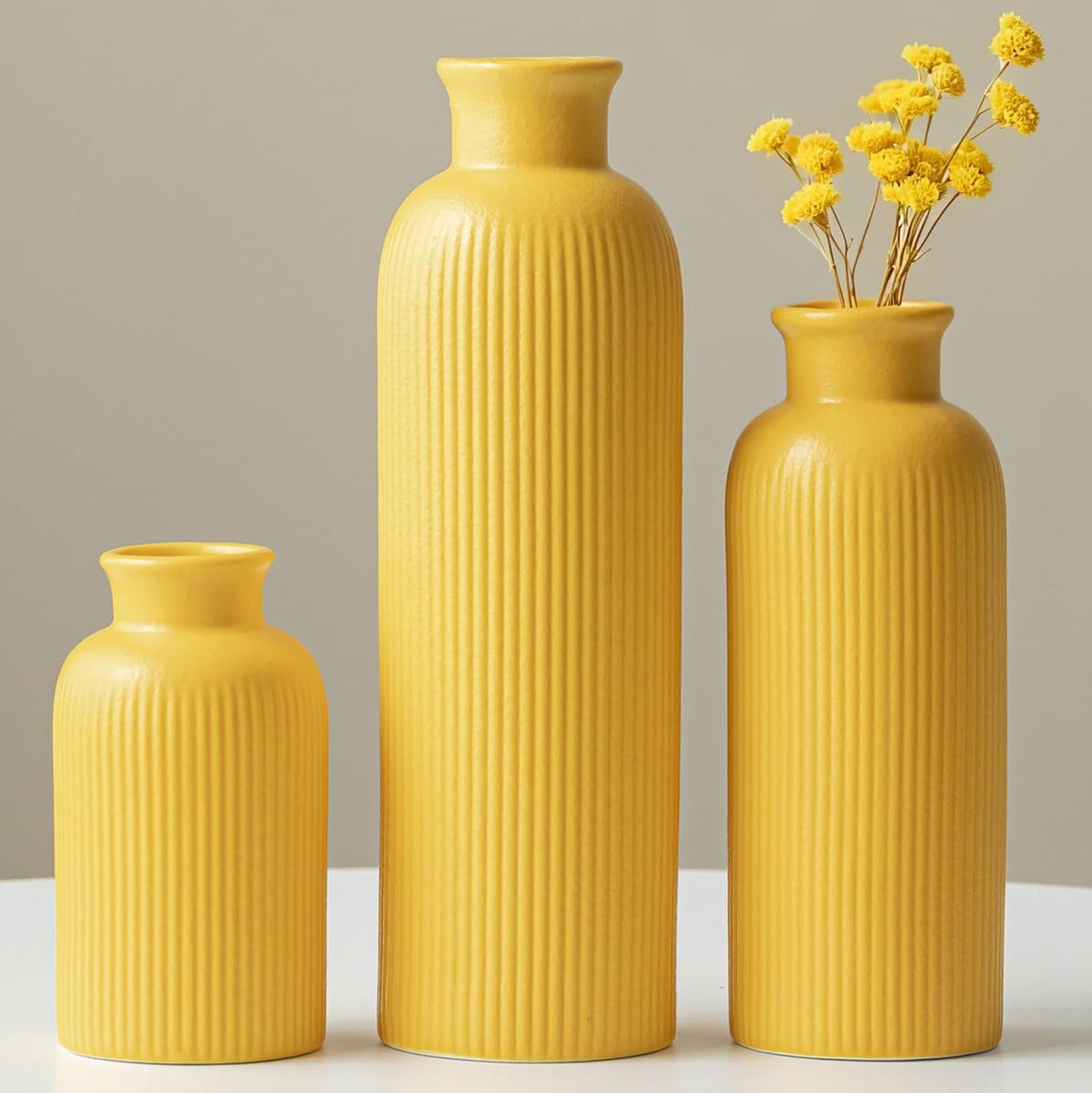 Three bright yellow ribbed ceramic vases on a white table, the small and tall right vase holding yellow dried flowers.