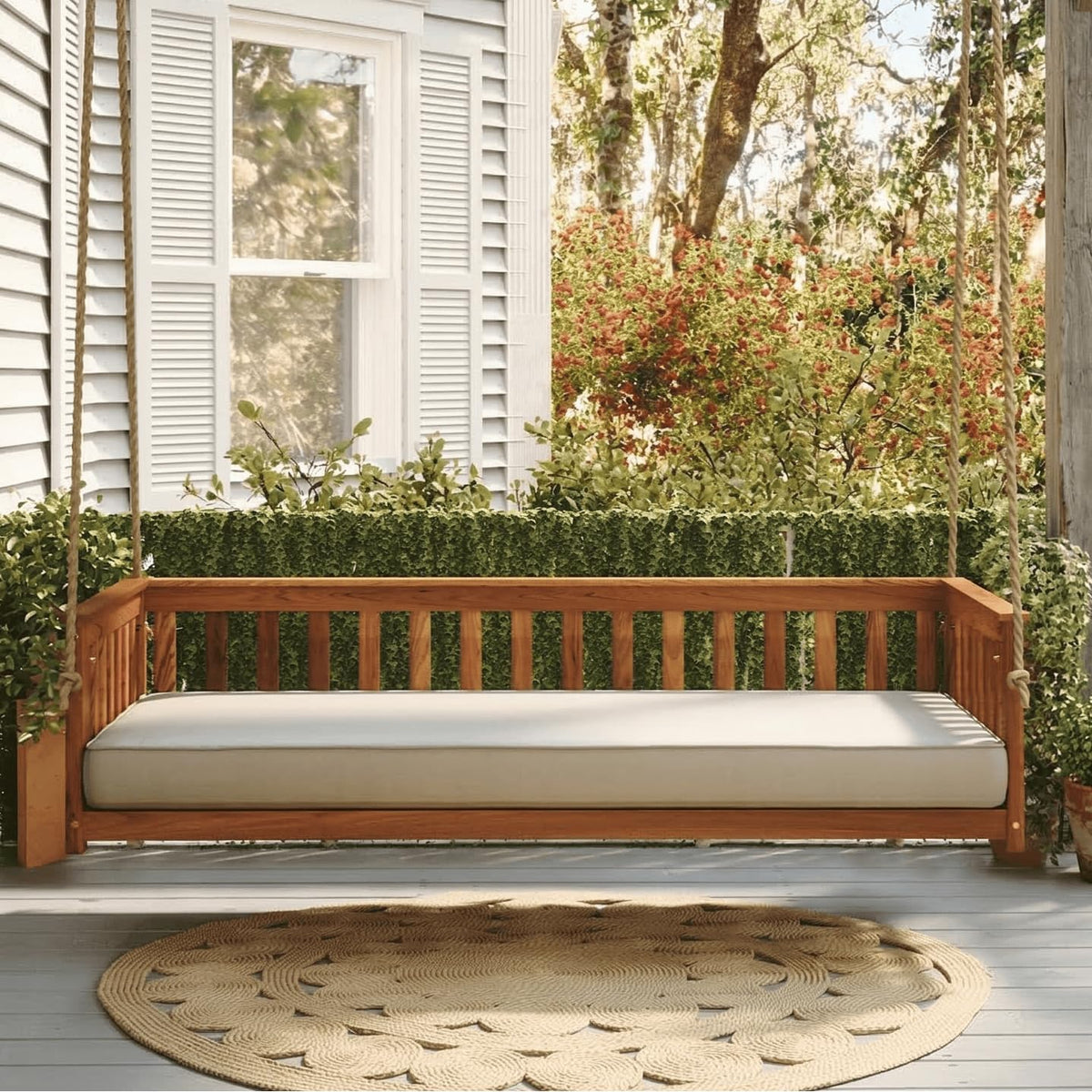 Porch scene with a hanging wooden daybed swing holding a light cushion beneath a window.