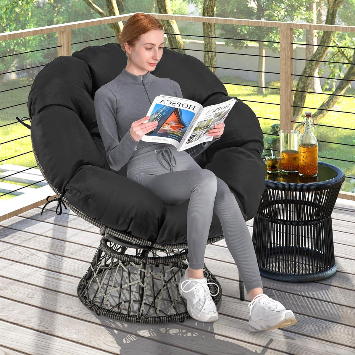 Person reading on the black papasan chair placed on a sunlit wooden deck beside a small round side table.