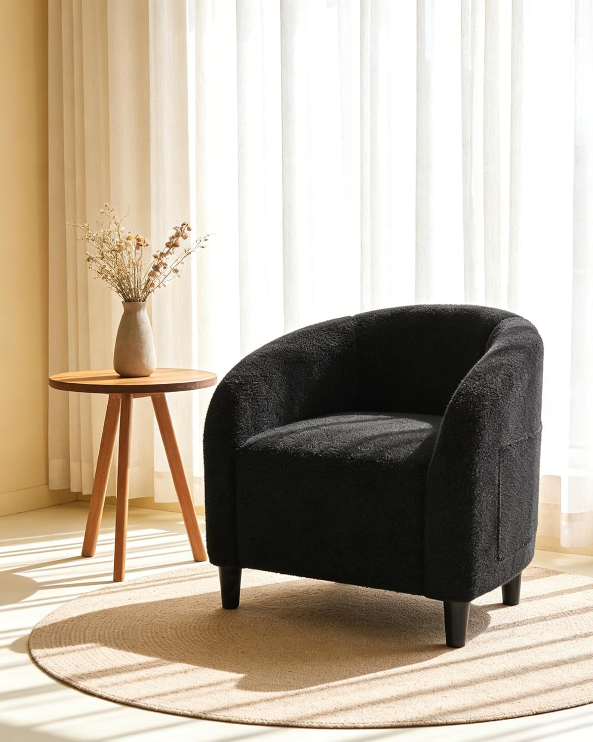 Black boucle barrel chair on round rug beside small wooden side table and vase by sheer curtains