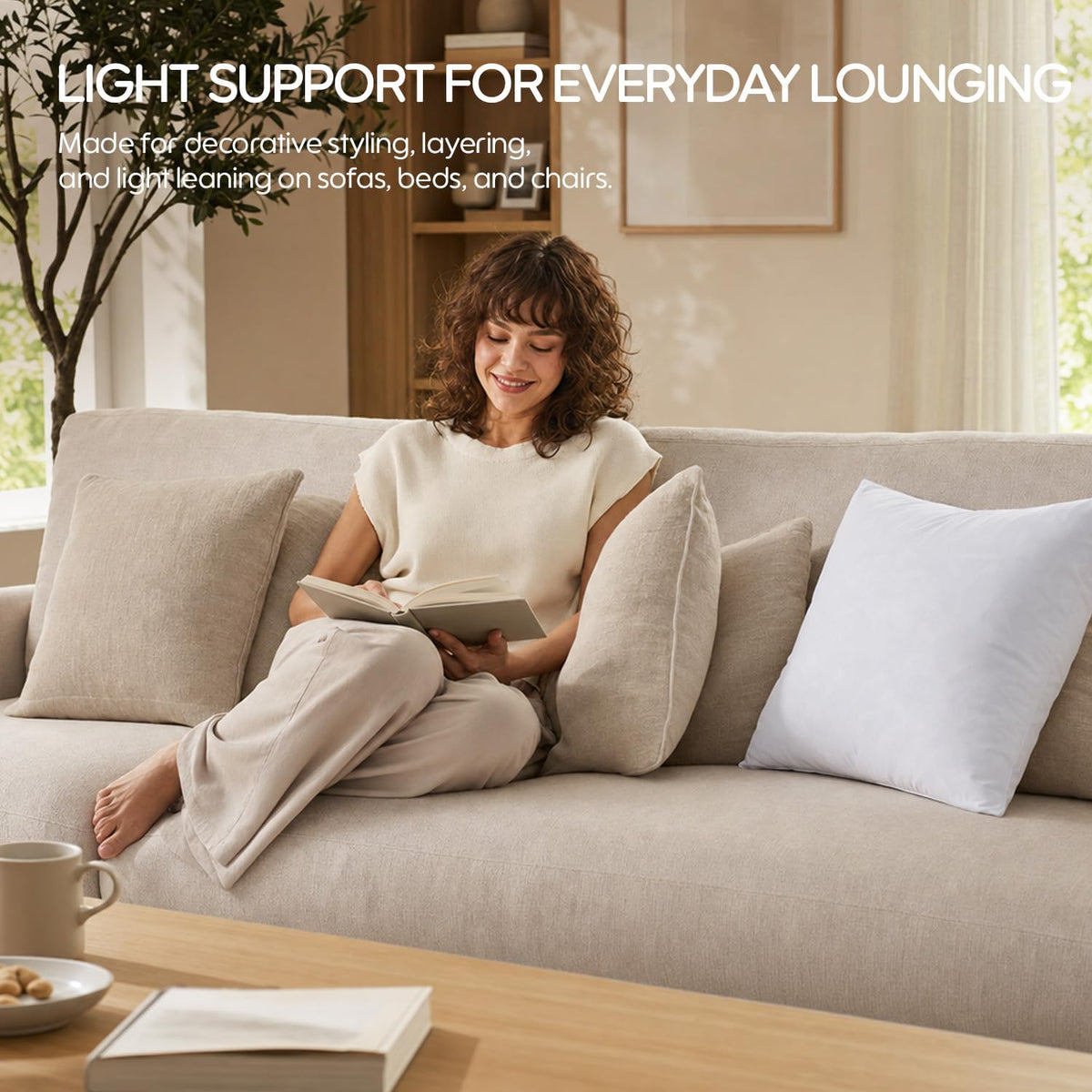 Person reading on a light sofa with beige pillows and a single plain white pillow insert propped at one end.