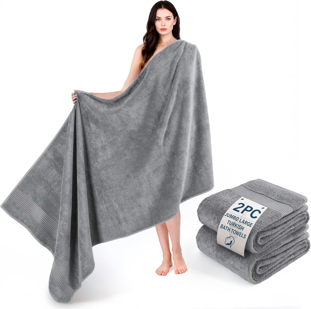 Woman holding and draped in a large gray bath towel with two folded gray towels beside her.