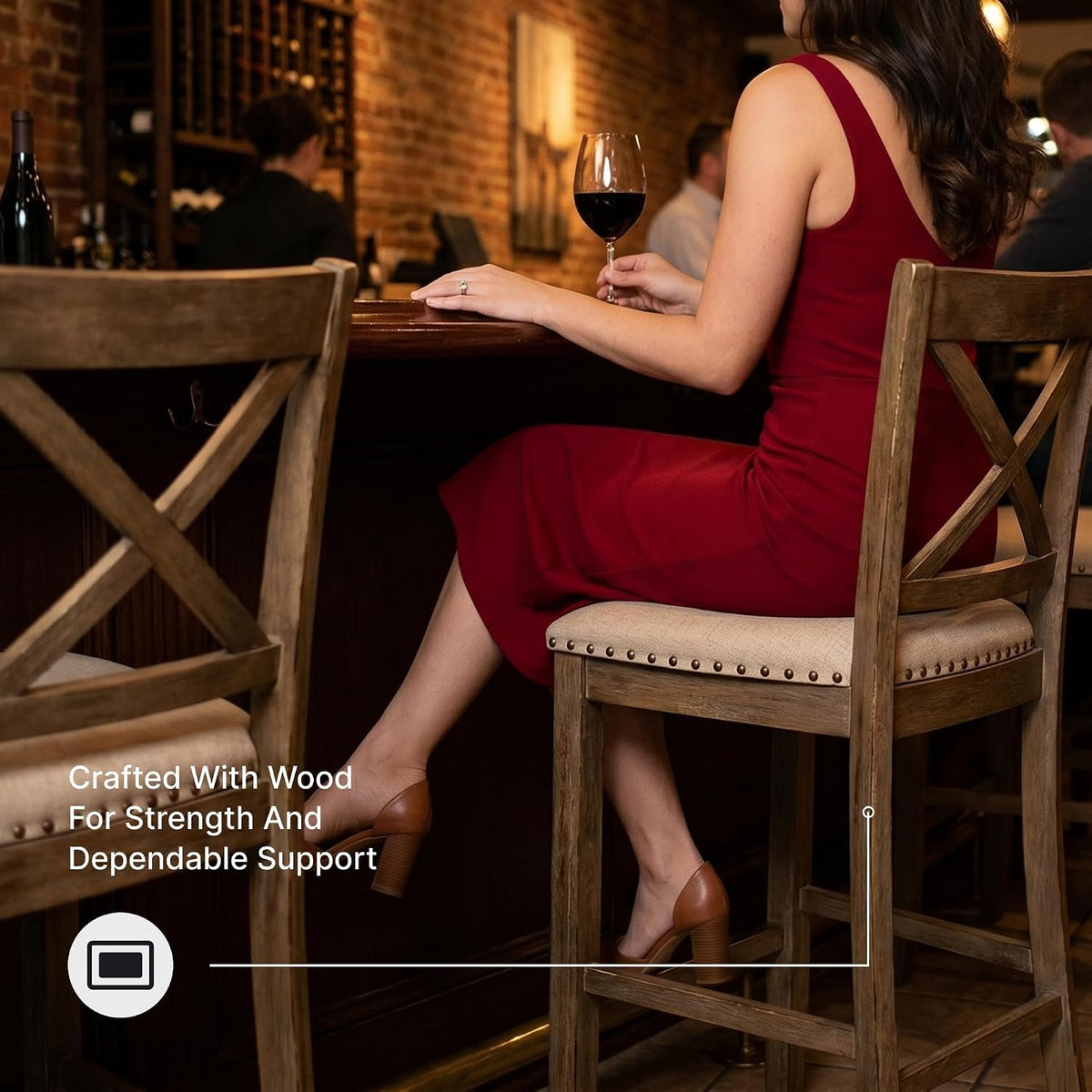 Woman seated on a beige upholstered counter stool at a dark wood bar, showing the X-back and nailhead trim