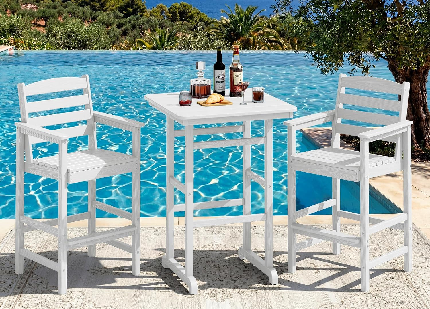 White bar table with two white armchairs arranged at a poolside rug, glasses and bottles on the table