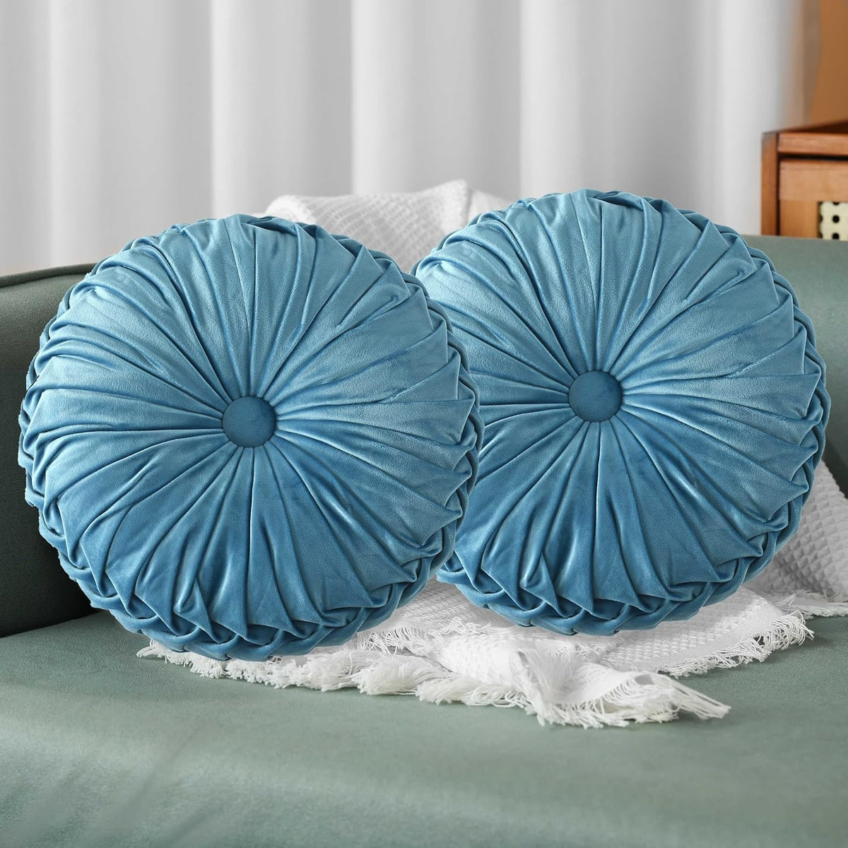 Pair of teal round ruched pillows with central button resting on a green sofa and white throw.