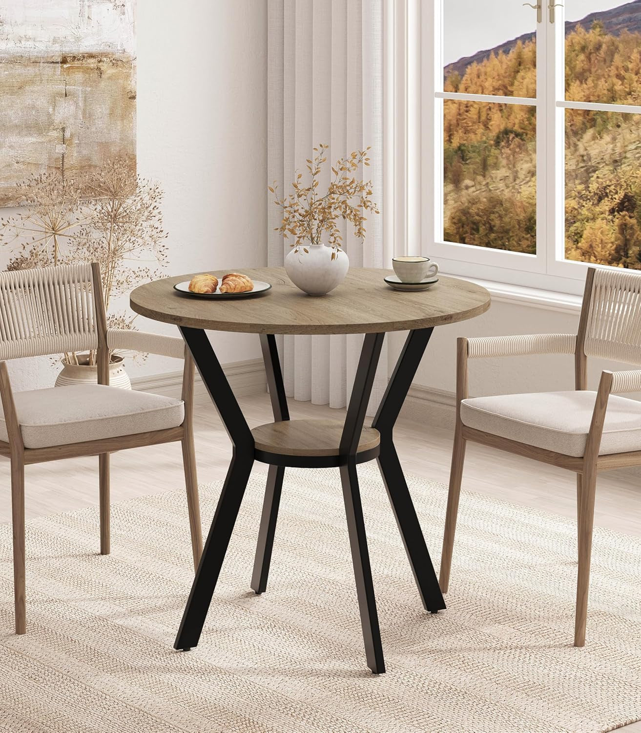 Round light brown table with black metal legs and small lower shelf, two woven-back chairs nearby.