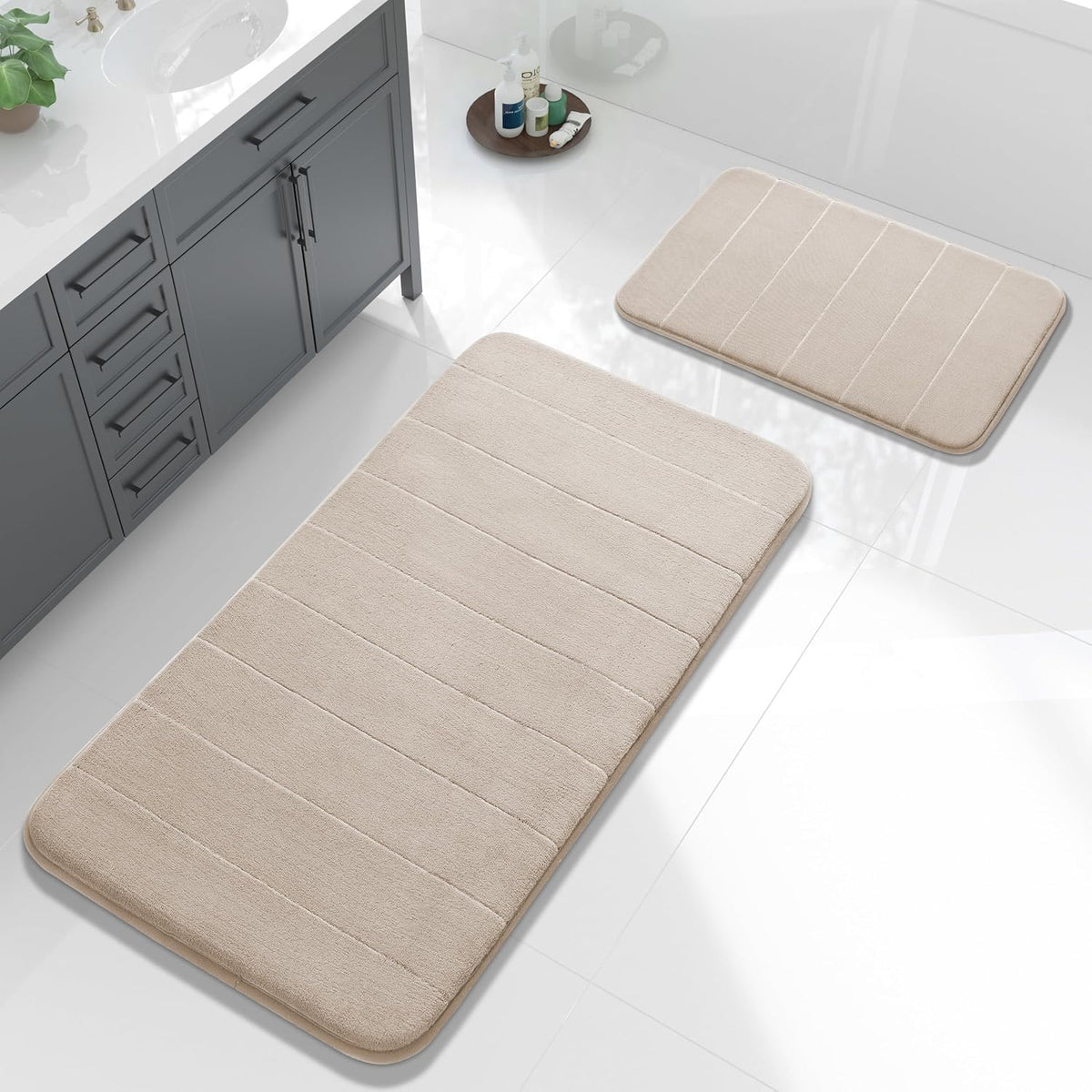 Light beige memory-foam bath mat set shown beside a sink and toiletries on white tile.
