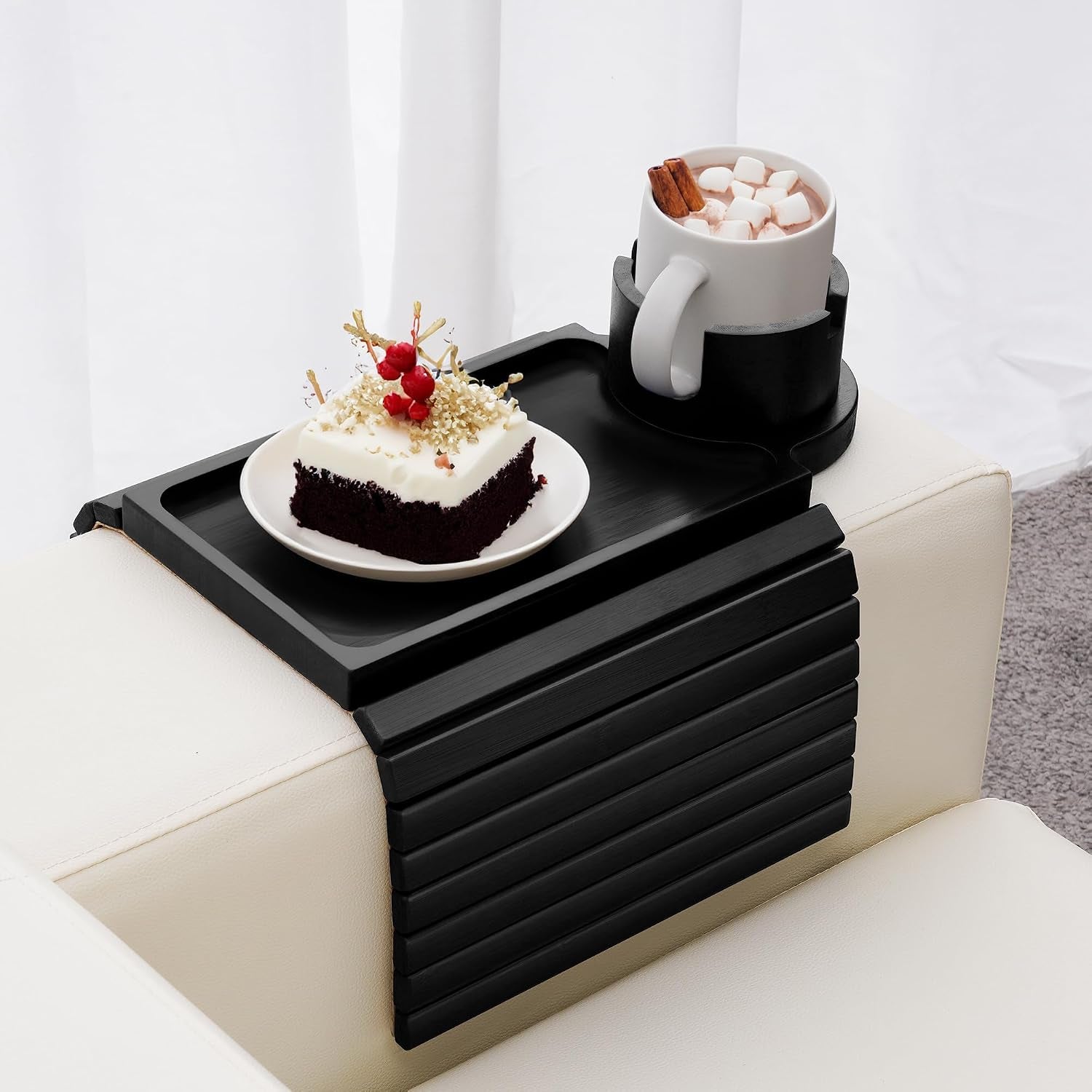 Black slatted bamboo arm tray draped over a cream sofa arm holding a plate with cake and a mug in the cup holder.