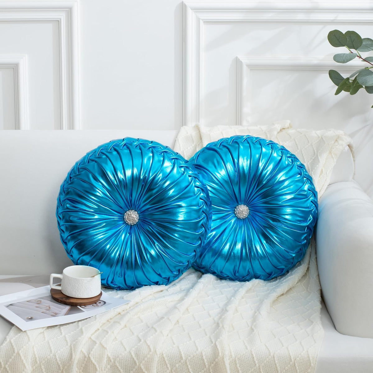 Shiny metallic blue round ruched pillows arranged on a white sofa with a knit throw