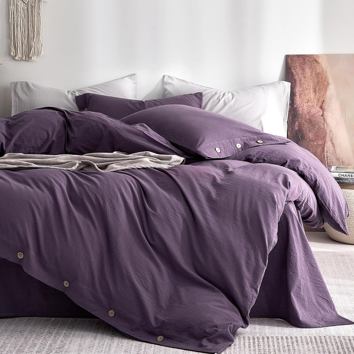 Close view of a rumpled purple duvet and pillowcases with wooden buttons against white pillows and a textured wall.