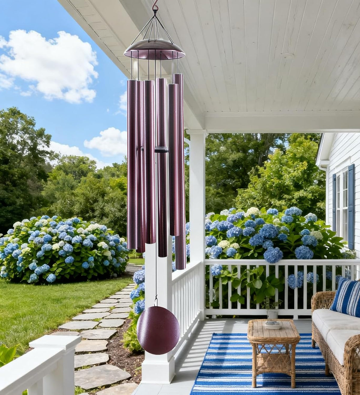 Wind chimes hanging from a white porch ceiling above a blue-striped rug and a bed of blue hydrangeas.