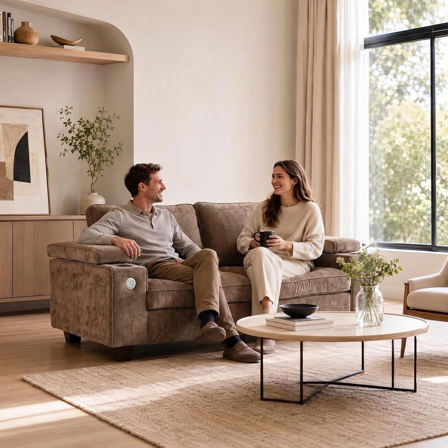 Man and woman sitting and smiling on a brown loveseat in a sunlit living room with a round coffee table.