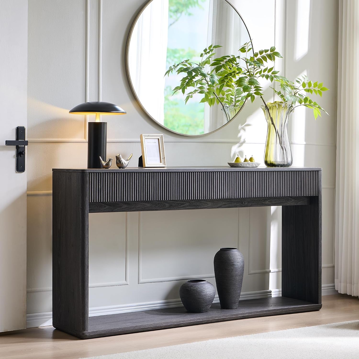 Angled, sunlit view of a dark gray fluted console with a lamp, round mirror and two black vases on the lower shelf.