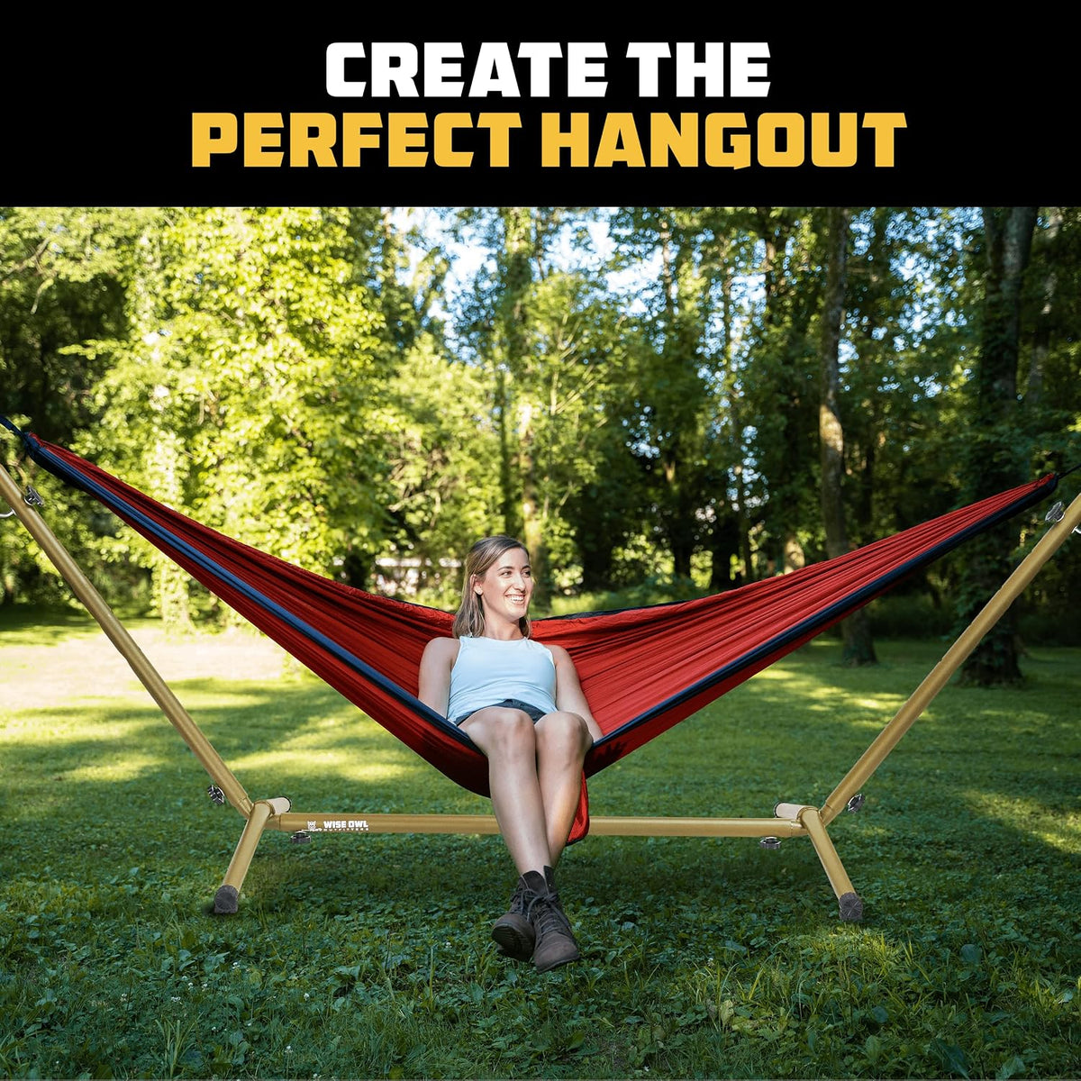 Woman sitting in a red hammock suspended on a gold steel stand in a grassy park.