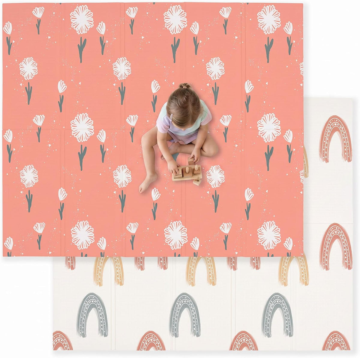 Overhead view of a child sitting on a coral play mat patterned with white daisies; rainbow arches show on the folded