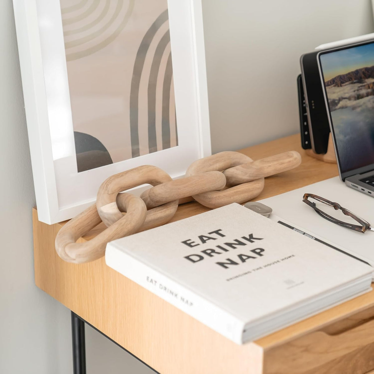 Close desk view of the acacia chain draped beside a framed print, glasses and a laptop on a light wood surface