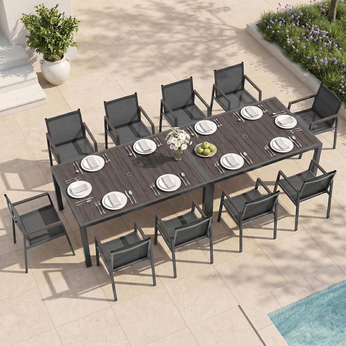 Long dark-wood outdoor table with multiple place settings and ten charcoal mesh chairs by a pool.