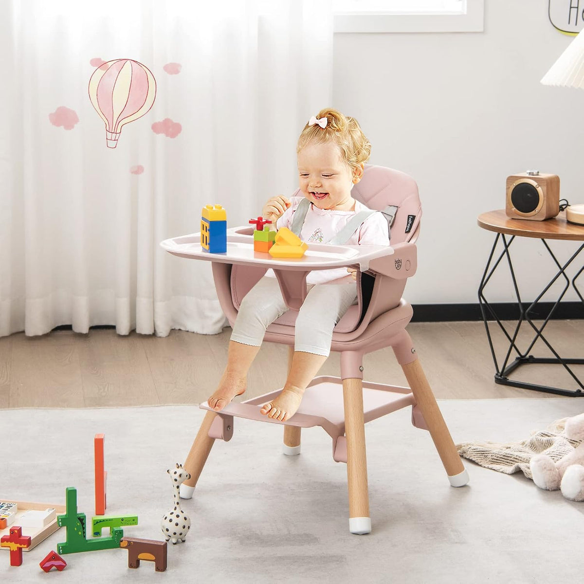 Toddler seated in a pink highchair with wooden legs and a detachable tray in a bright nursery.