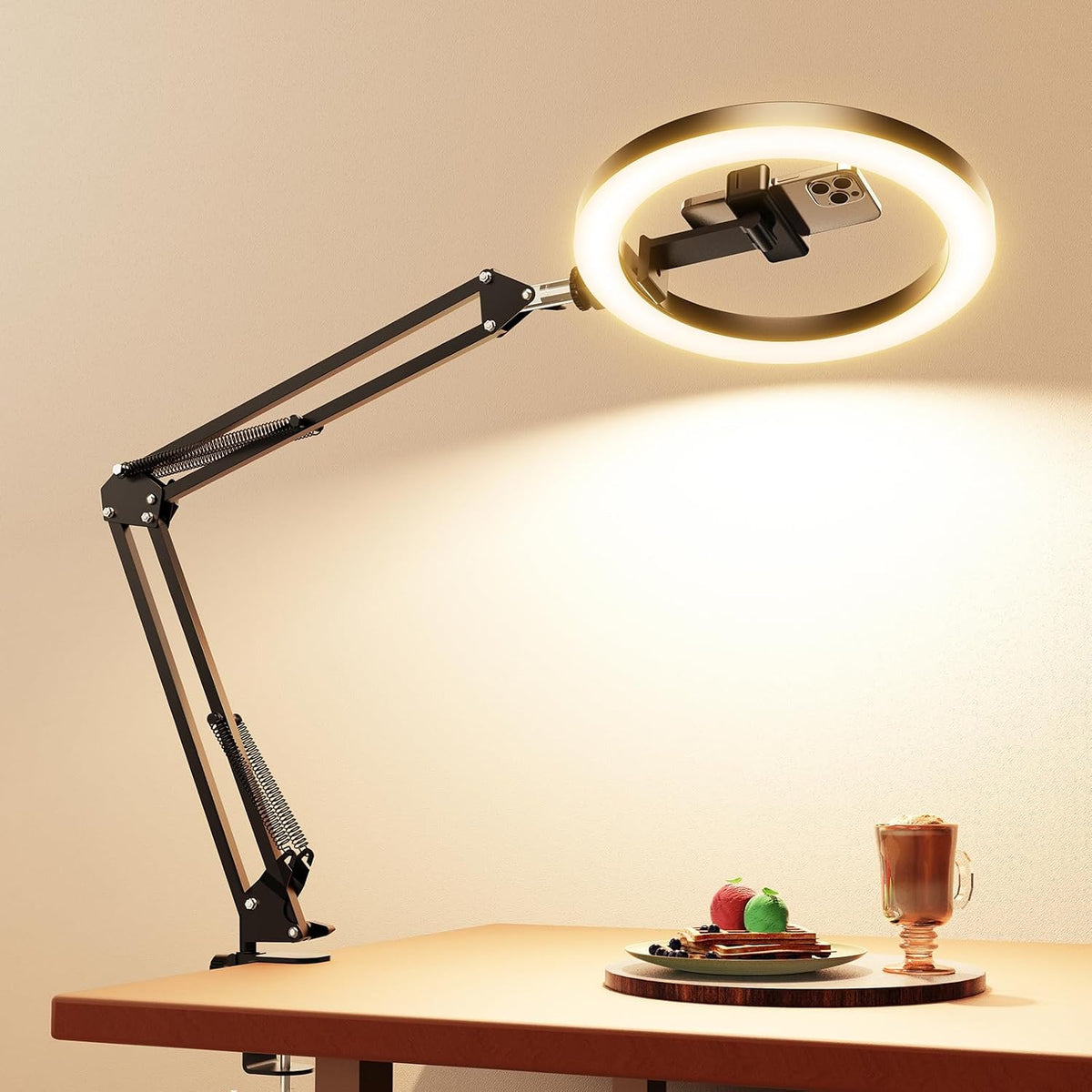 Black clamp-mounted desk arm with illuminated round ring light angled over a wooden tabletop with plated food and a drink