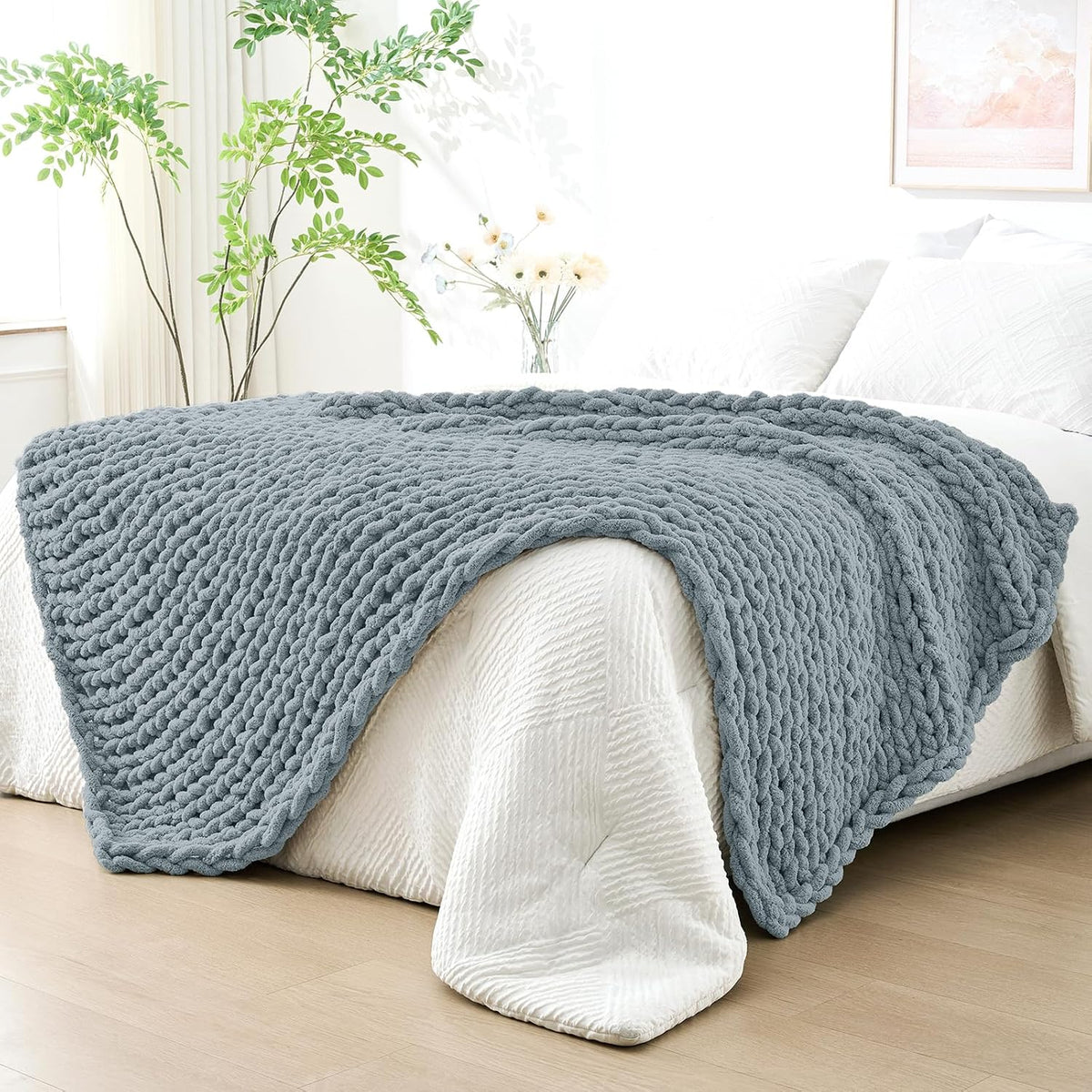Blue-gray chunky knit throw draped over the foot of a white bed in a bright bedroom.