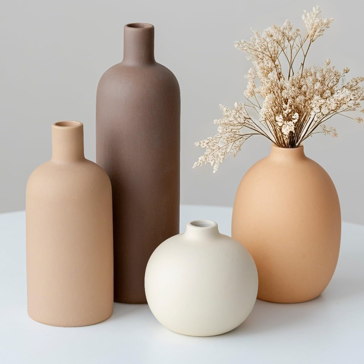 Front view of four matte ceramic vases on a white table: small cream round vase front, tall brown bottle behind, beige left