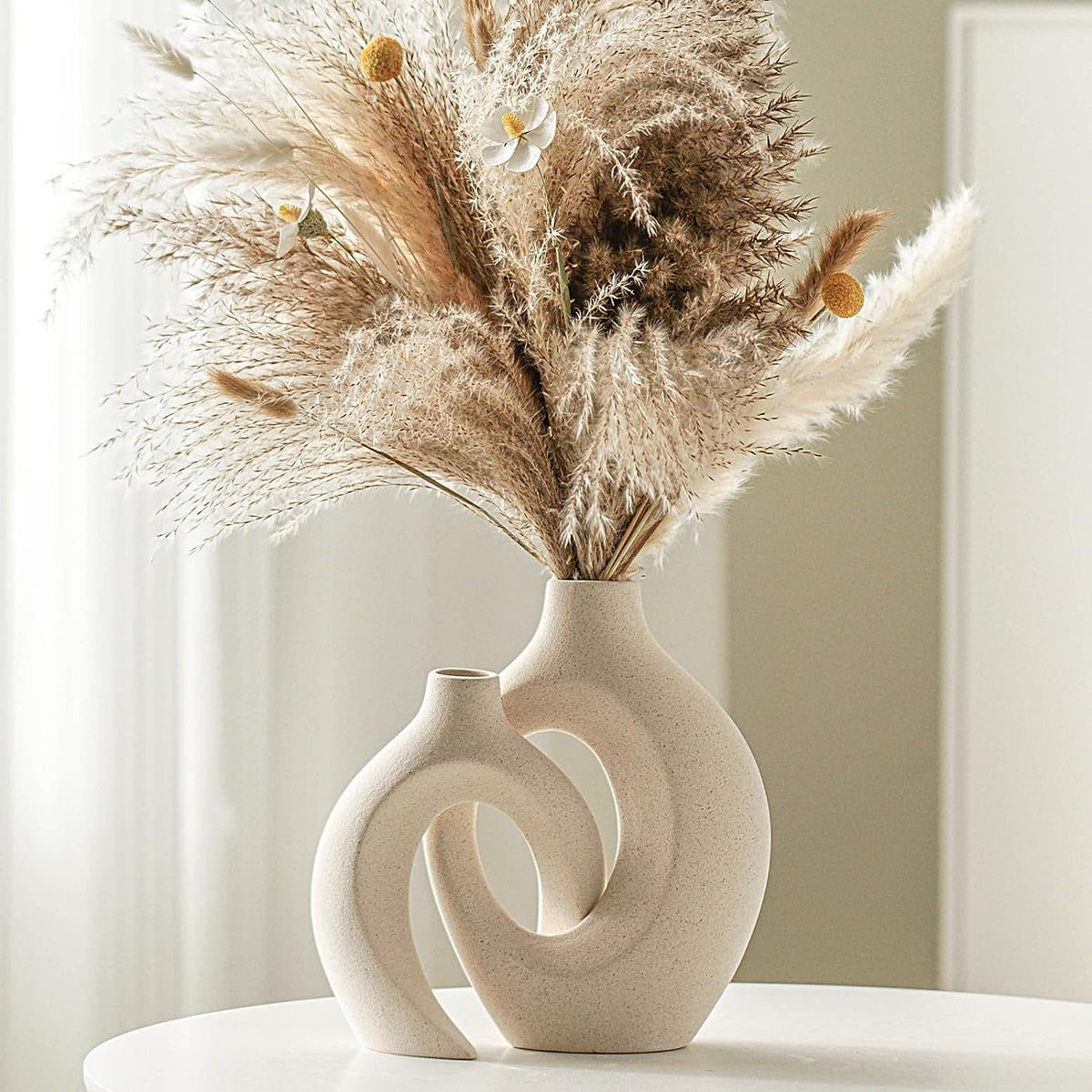Matte beige ceramic sculptural vase pair displayed with pampas grass on a white round table