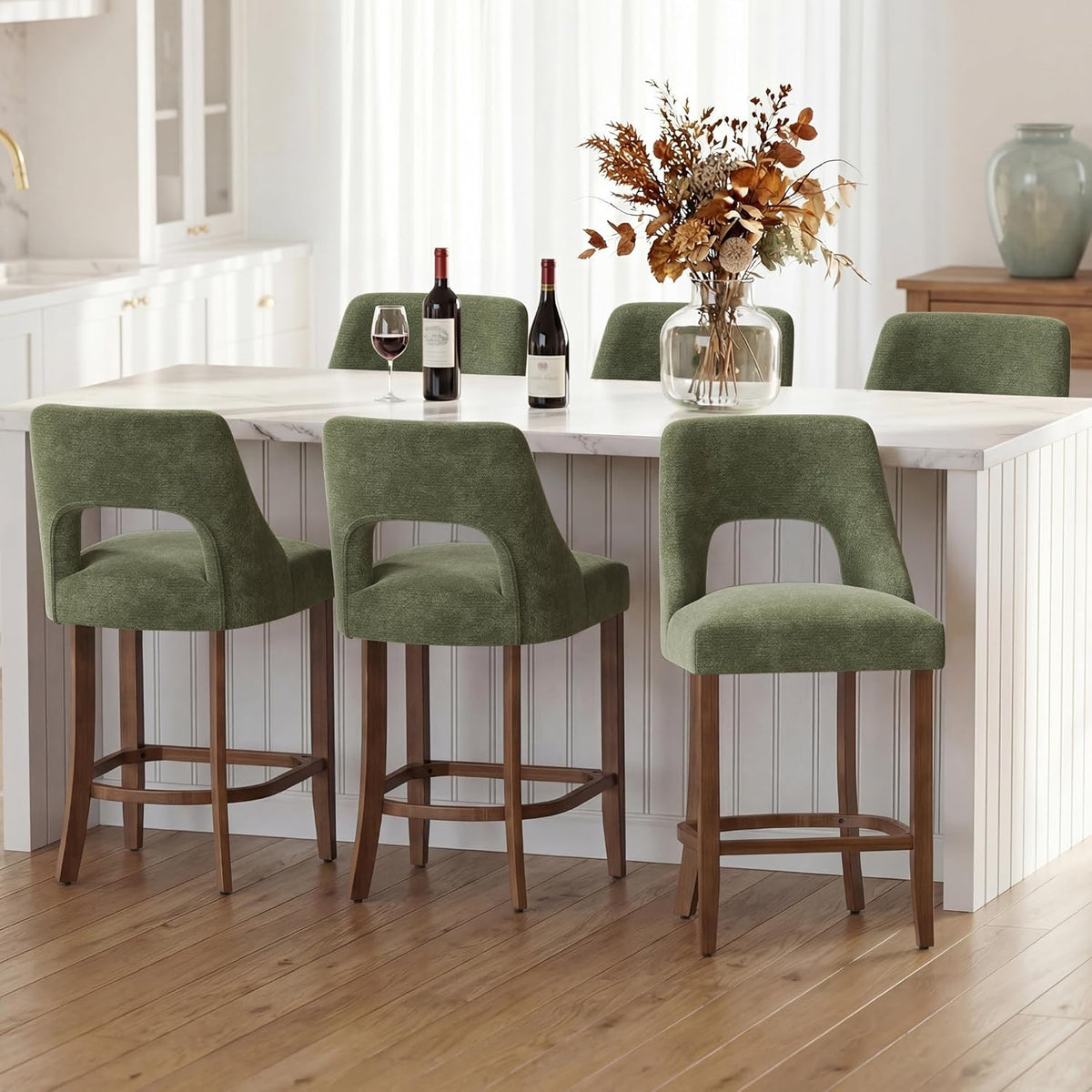 Three green upholstered counter stools lined at a kitchen island with wine bottles and a vase on top.