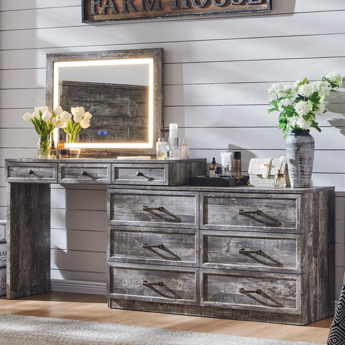 Rustic gray wood dresser with eight drawers beneath a lit framed mirror and vases with flowers on the surface.