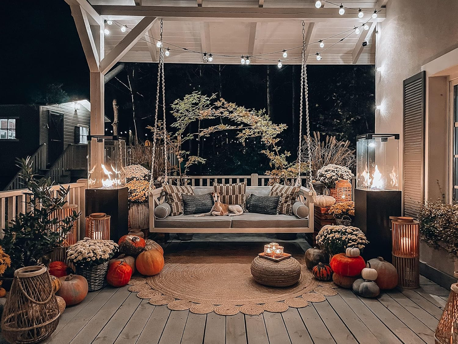 Cozy covered porch at night with a swing sofa flanked by two lit glass patio heaters and autumn decor.