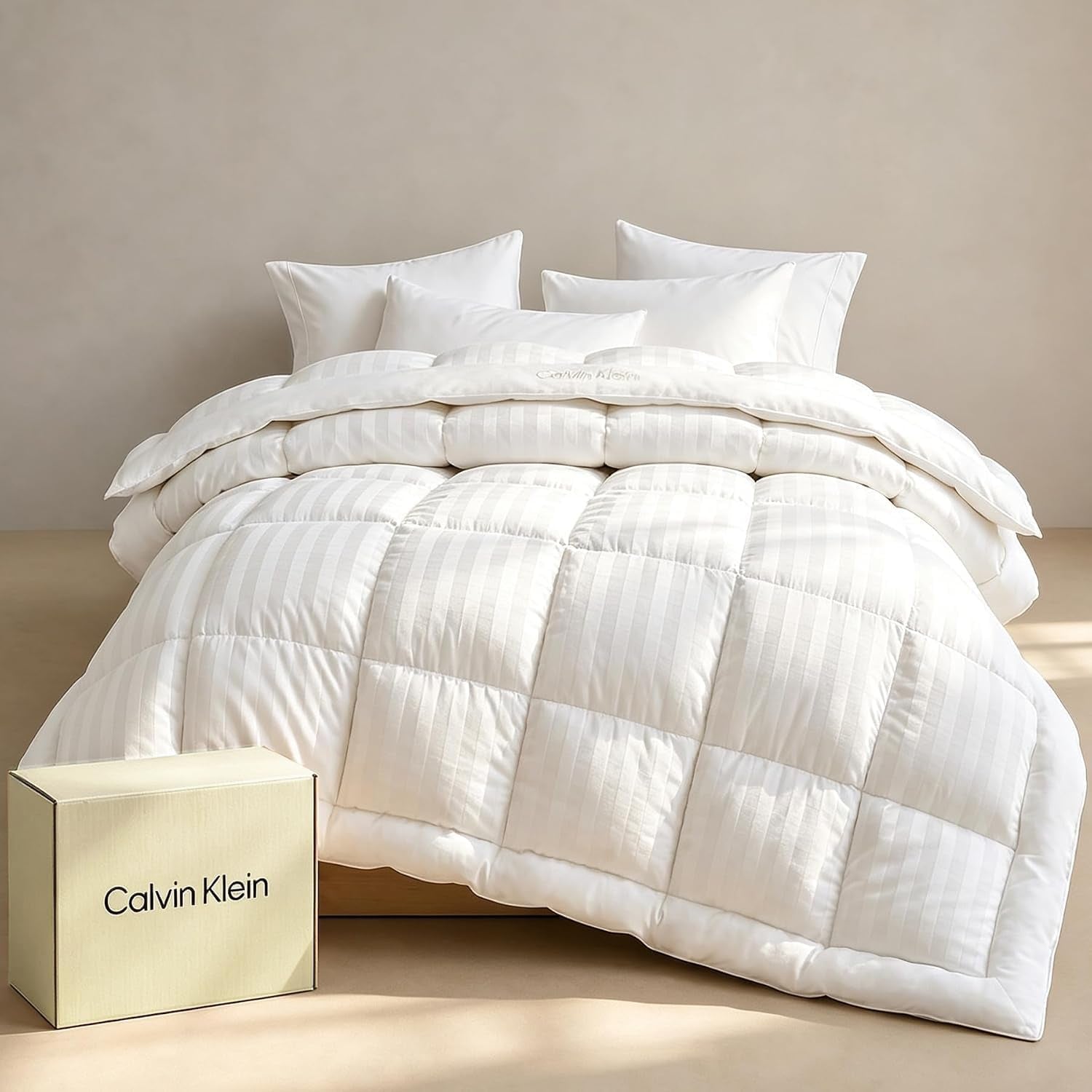 White striped box-stitched comforter draped on bed with stacked pillows and a beige packaging box at the floor