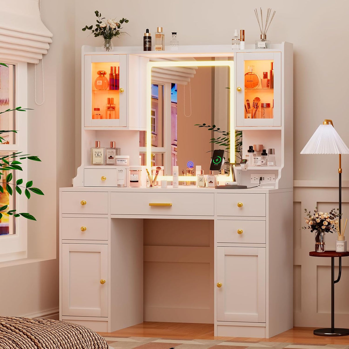 White vanity desk with LED mirror and glass-front cabinets lit orange, tabletop cosmetics and phone charger on the desktop