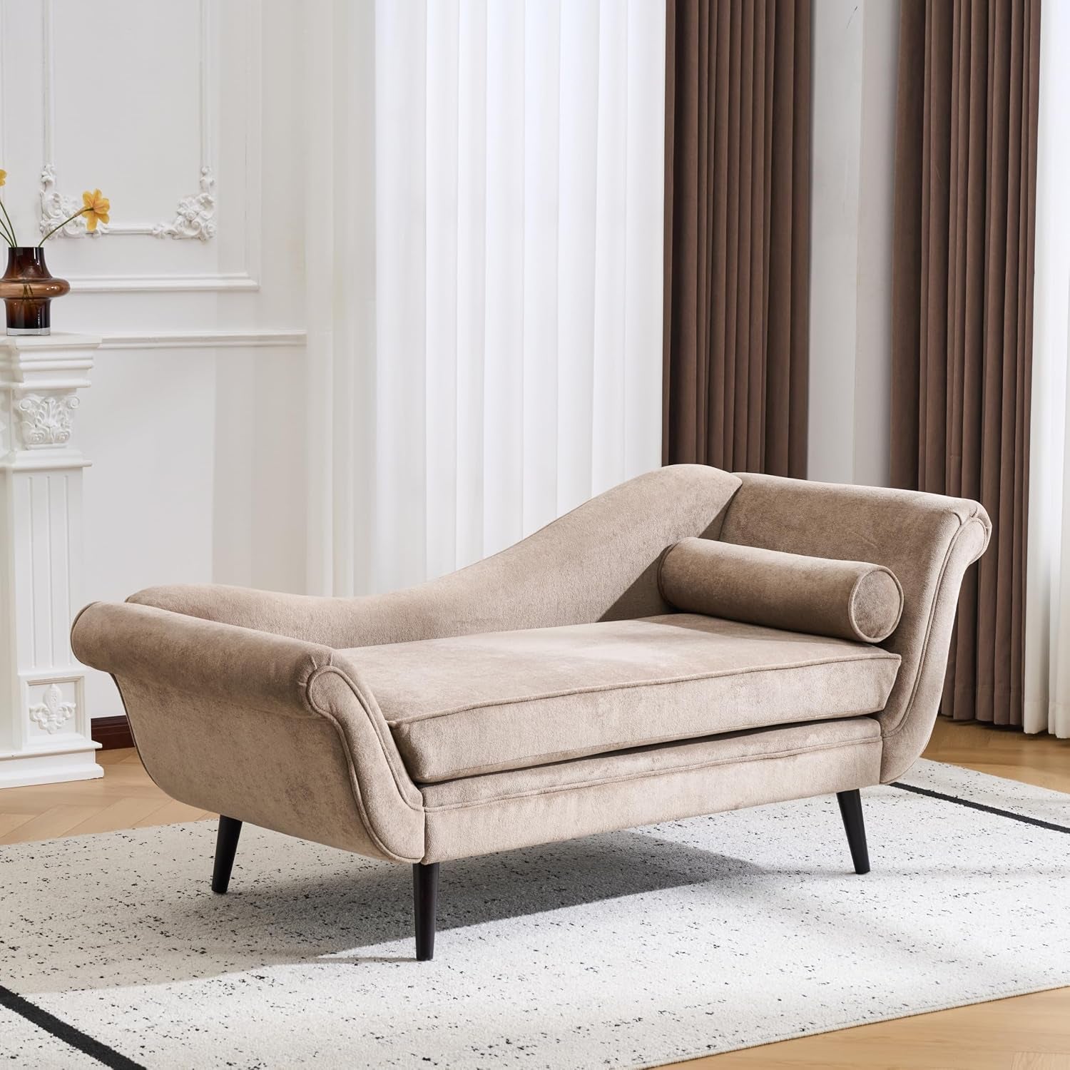 Light-brown velvet chaise lounge in a bright living room on a patterned rug with curtains behind