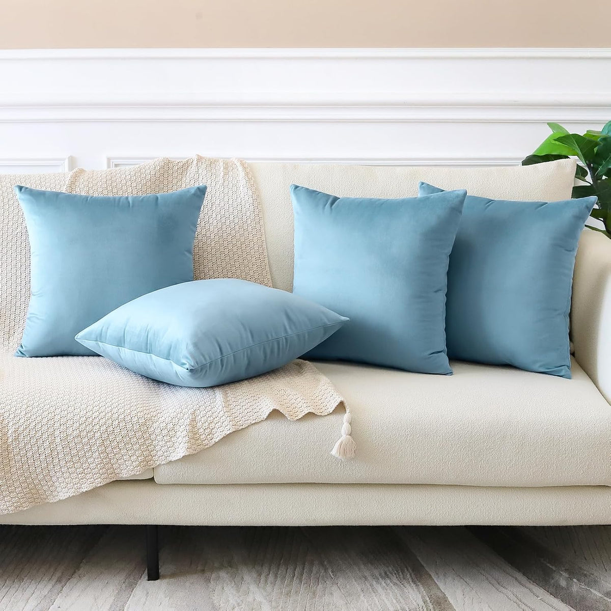 Three light-blue square pillows and one laying flat on a cream sofa draped with a knitted throw
