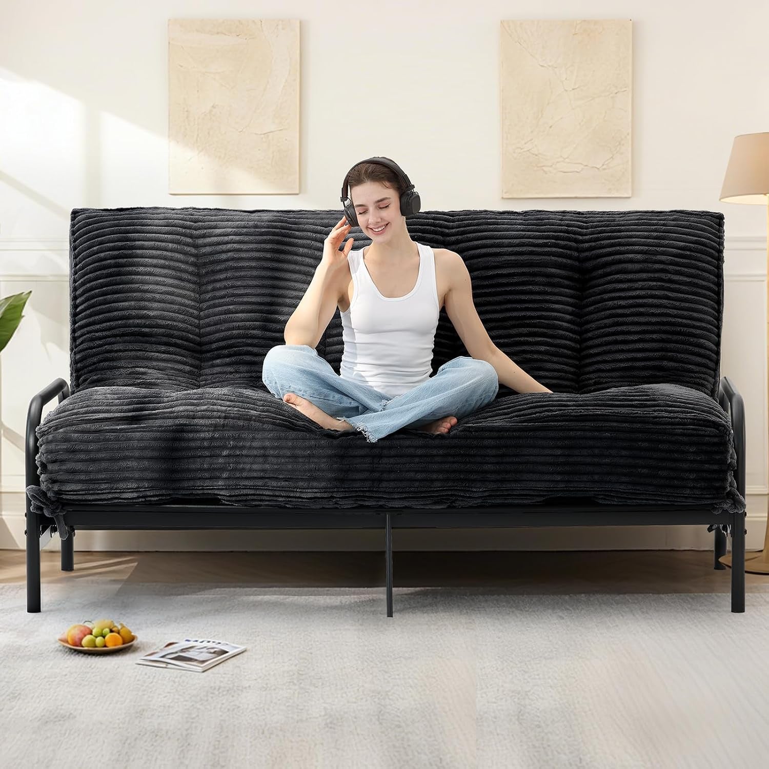Front view of grey futon on a black metal frame with a seated woman wearing headphones on the cushion.